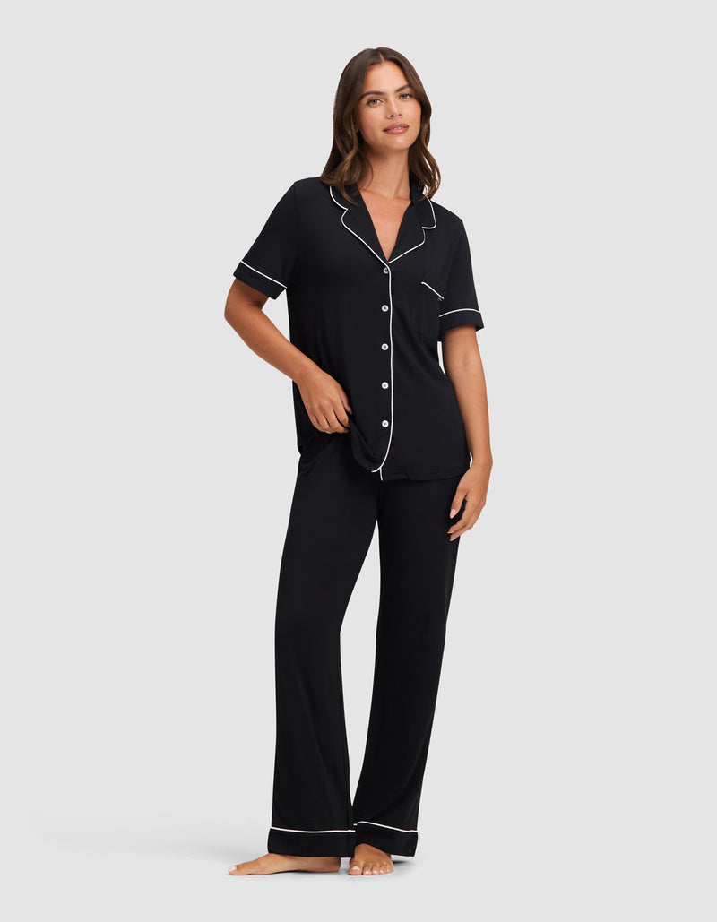 A woman stands barefoot against a light gray background, wearing Cozy Earth’s Women's Bamboo Stretch-Knit Short Sleeve & Pant Pajama Set in black with white piping. The set includes a short-sleeved button-up top and long pants.