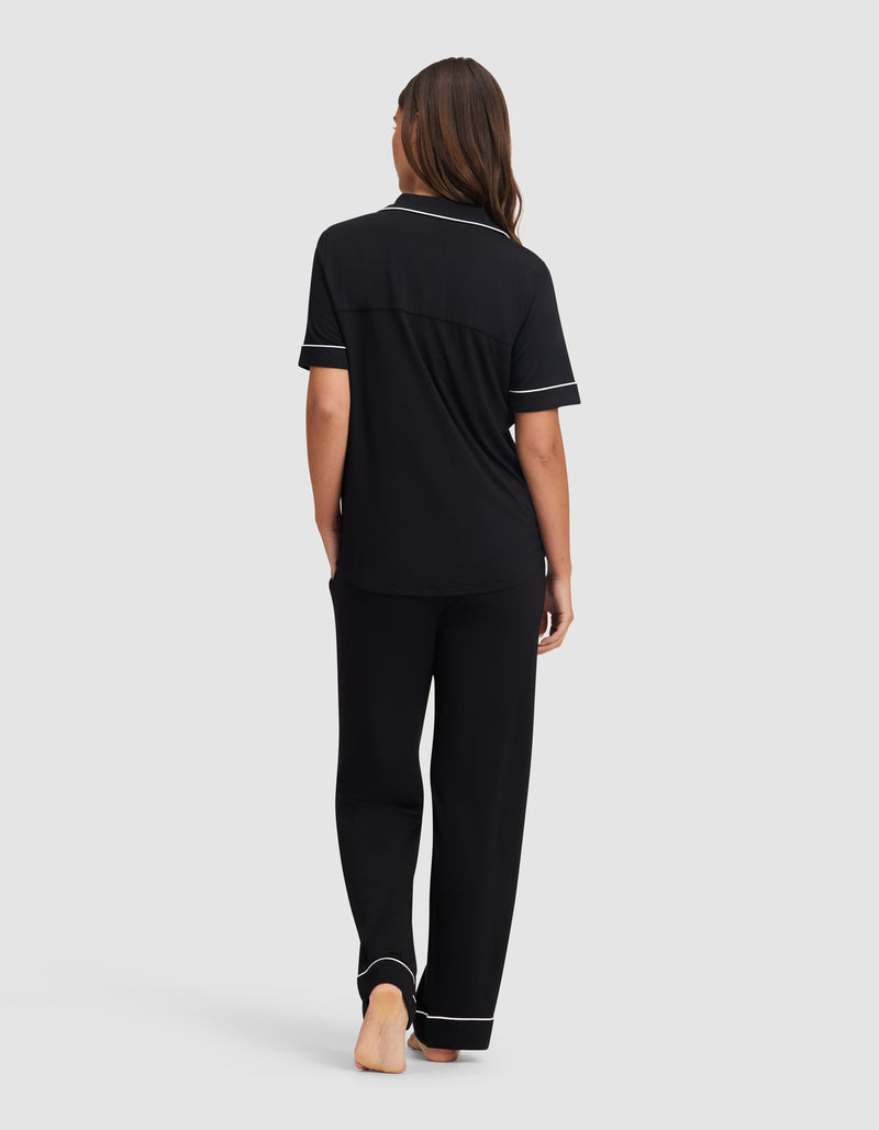 A woman stands barefoot facing away, wearing the Cozy Earth Women's Bamboo Stretch-Knit Short Sleeve & Pant Pajama Set in black with white piping, set against a plain light gray background.