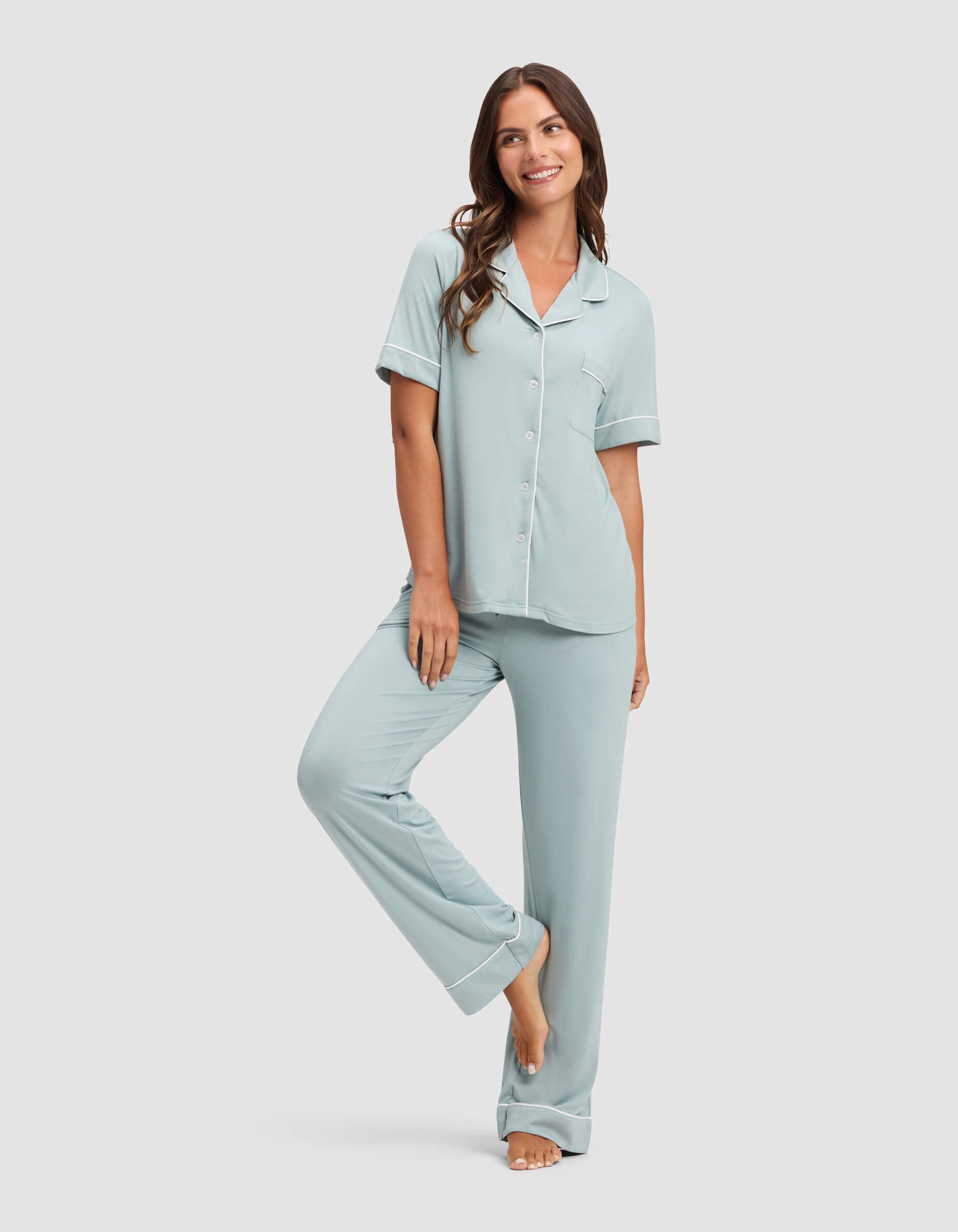 A woman smiles barefoot, one leg raised, modeling Cozy Earth's Women's Bamboo Stretch-Knit Pajama Pant in light blue with a short-sleeve button-up top, against a plain light background.|Color:Brook