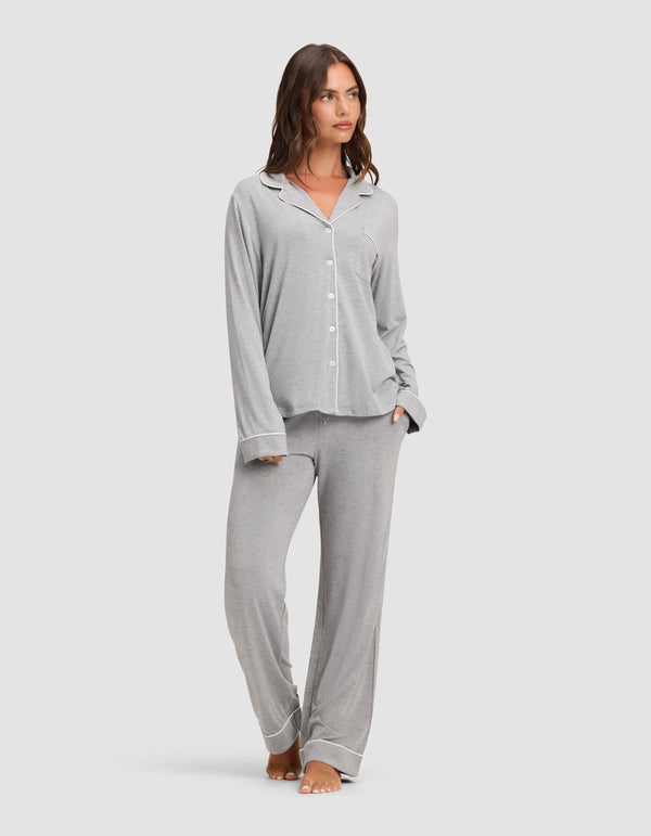 A woman stands barefoot against a plain light background, wearing the Cozy Earth Women's Bamboo Stretch-Knit Long Sleeve Pajama Set in light grey with white piping, featuring a button-up top and matching pants.  |Color:Grey