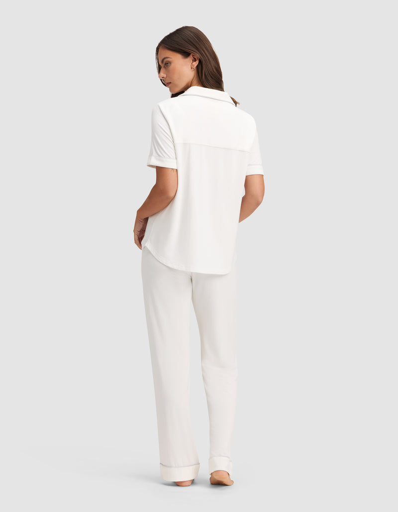 A woman faces away, barefoot on a light gray background, wearing Cozy Earth's Women's Bamboo Stretch-Knit Short Sleeve & Pant Pajama Set in white.