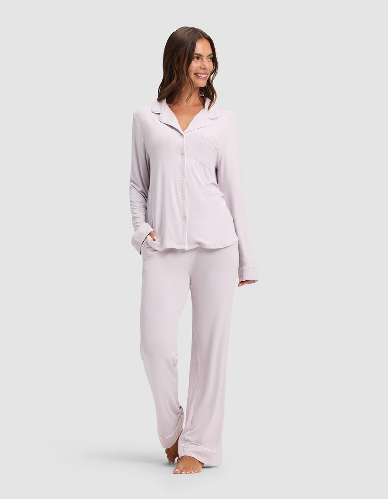A woman smiles while wearing the Cozy Earth Women's Bamboo Stretch-Knit Long Sleeve Pajama Set, featuring a button-up top and matching pants in light lavender, standing barefoot against a plain light background.