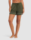 A woman stands barefoot on a light background, hands in the pockets of HIDE Women's Bamboo Stretch-Knit Pajama Shorts in olive green with white trim. |Color:Olive