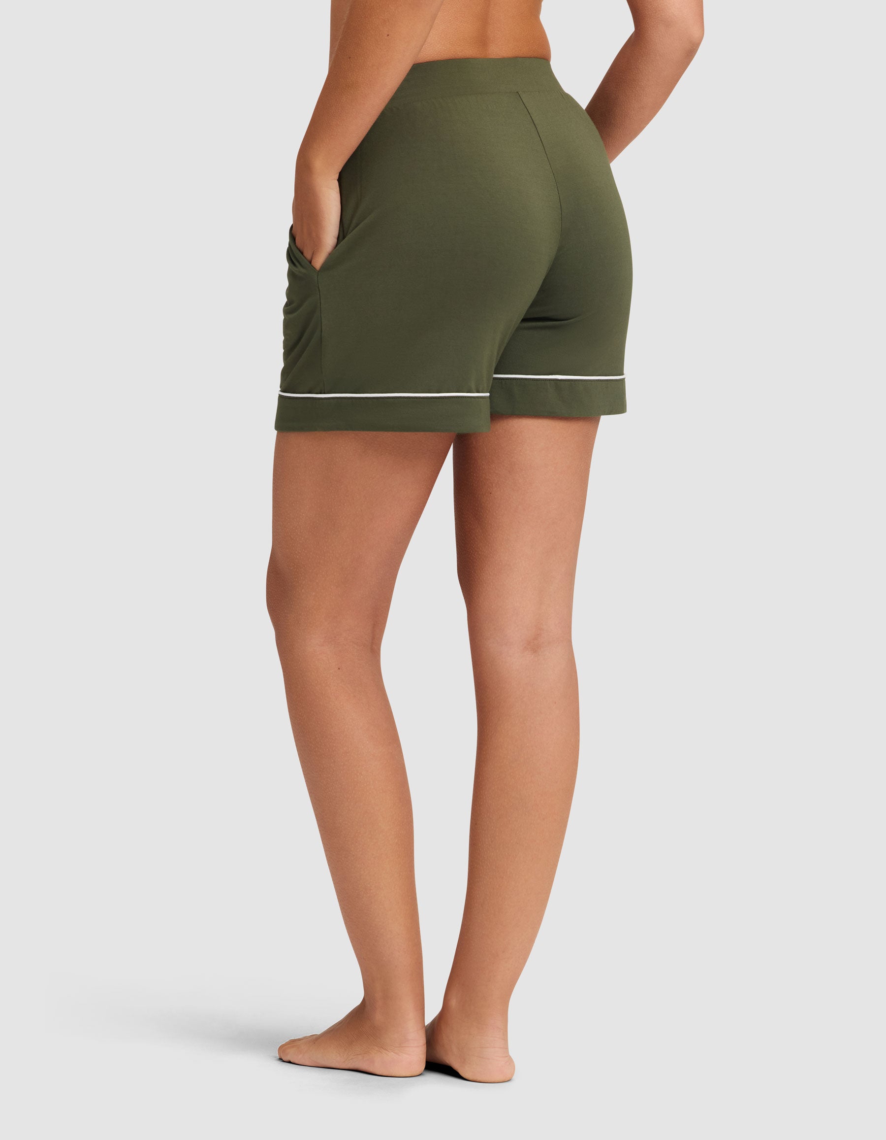 A woman models HIDE's Women's Bamboo Stretch-Knit Pajama Short in olive green with white piping, standing barefoot on a white background, viewed from the back with one hand in a pocket. |Color:Olive