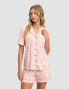 A woman with long, wavy blonde hair models the Cozy Earth Women's Bamboo Stretch-Knit Short Sleeve Pajama Top and matching shorts in a peach and white floral print, standing against a plain gray background.|Color:Melon Toile