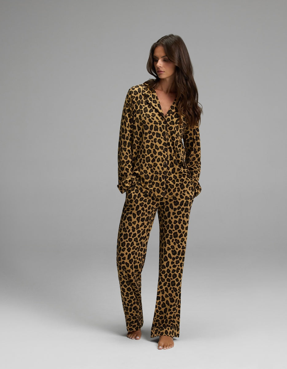 A woman stands barefoot against a plain gray background, wearing Cozy Earth’s Women’s Bamboo Stretch-Knit Long Sleeve Pajama Top and matching pants in a brown and black leopard print pattern. Her hands are in her pockets as she looks to the side.