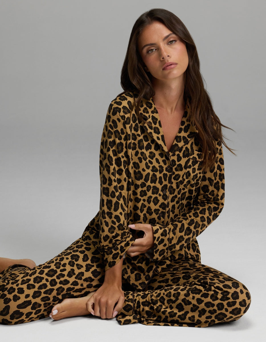 A woman with long brown hair sits on the floor against a plain gray background, wearing a matching set featuring the Cozy Earth Women’s Bamboo Stretch-Knit Long Sleeve Pajama Top in leopard print. She looks at the camera with a relaxed, neutral expression.