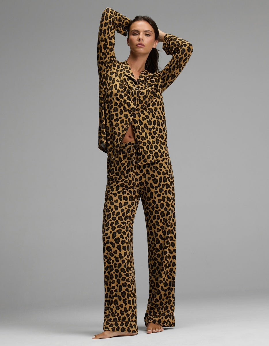 A woman poses barefoot against a plain gray background, wearing the Cozy Earth Women’s Bamboo Stretch-Knit Long Sleeve Pajama Set featuring a matching top and wide-leg pants in leopard print.