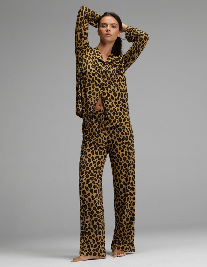 A woman poses barefoot against a plain gray background, wearing the Cozy Earth Women’s Bamboo Stretch-Knit Long Sleeve Pajama Set featuring a matching top and wide-leg pants in leopard print.