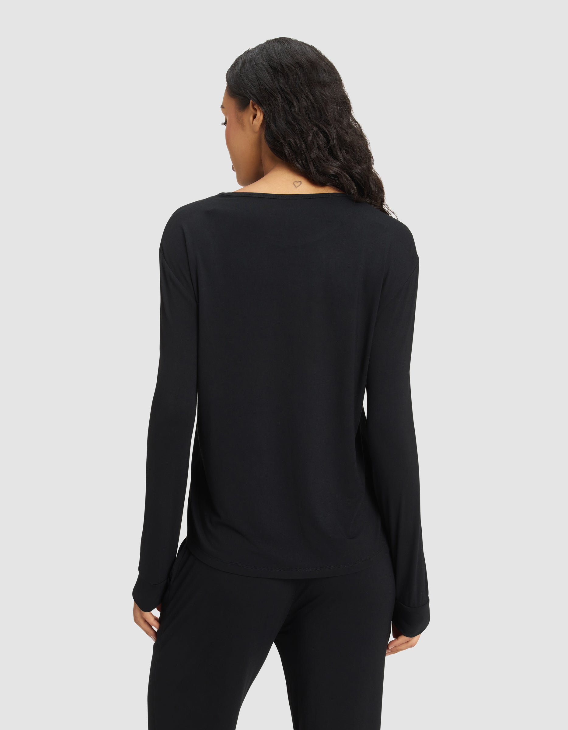 A woman with long dark hair is seen from behind wearing the Cozy Earth Women's Bamboo Stretch-Knit Long Sleeve Lounge Tee in black, paired with matching pants, standing against a plain light gray background. |Color:Black