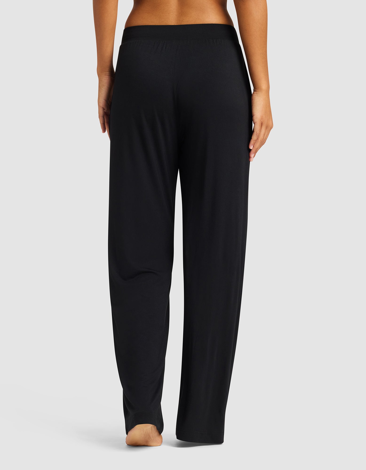 A person stands barefoot, facing away, wearing Cozy Earth’s Women's Bamboo Stretch-Knit Pant in black with an elastic waistband, set against a plain light gray background.|Color:Black