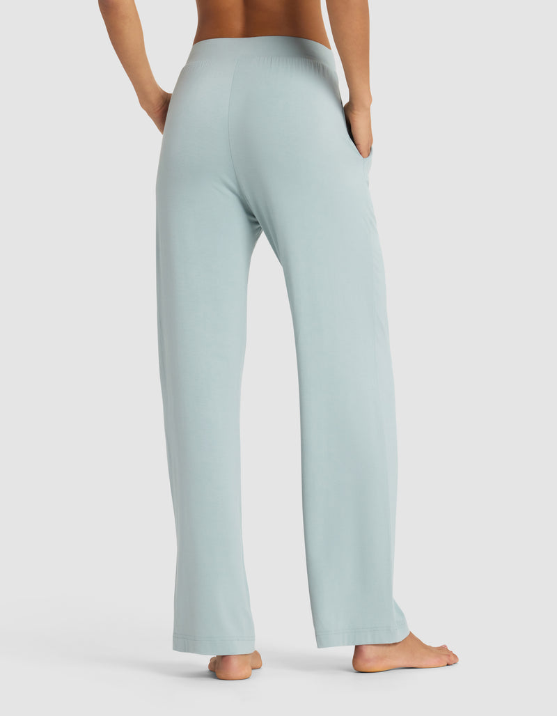 A woman stands barefoot, viewed from behind, wearing Cozy Earth’s Women's Bamboo Stretch-Knit Pant in light blue with hands in the side pockets. The background is plain white.