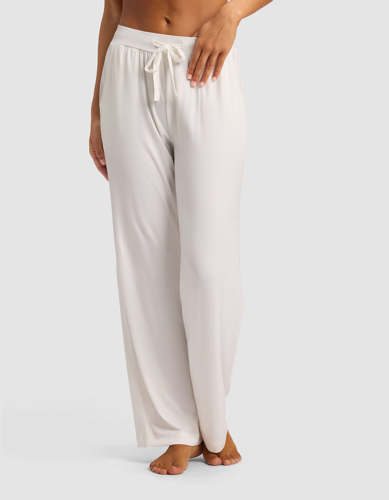 A woman stands barefoot against a plain light background, modeling the lower half of Cozy Earth's Women's Bamboo Stretch-Knit Pant—loose-fitting, light-colored lounge pants with a drawstring waist.