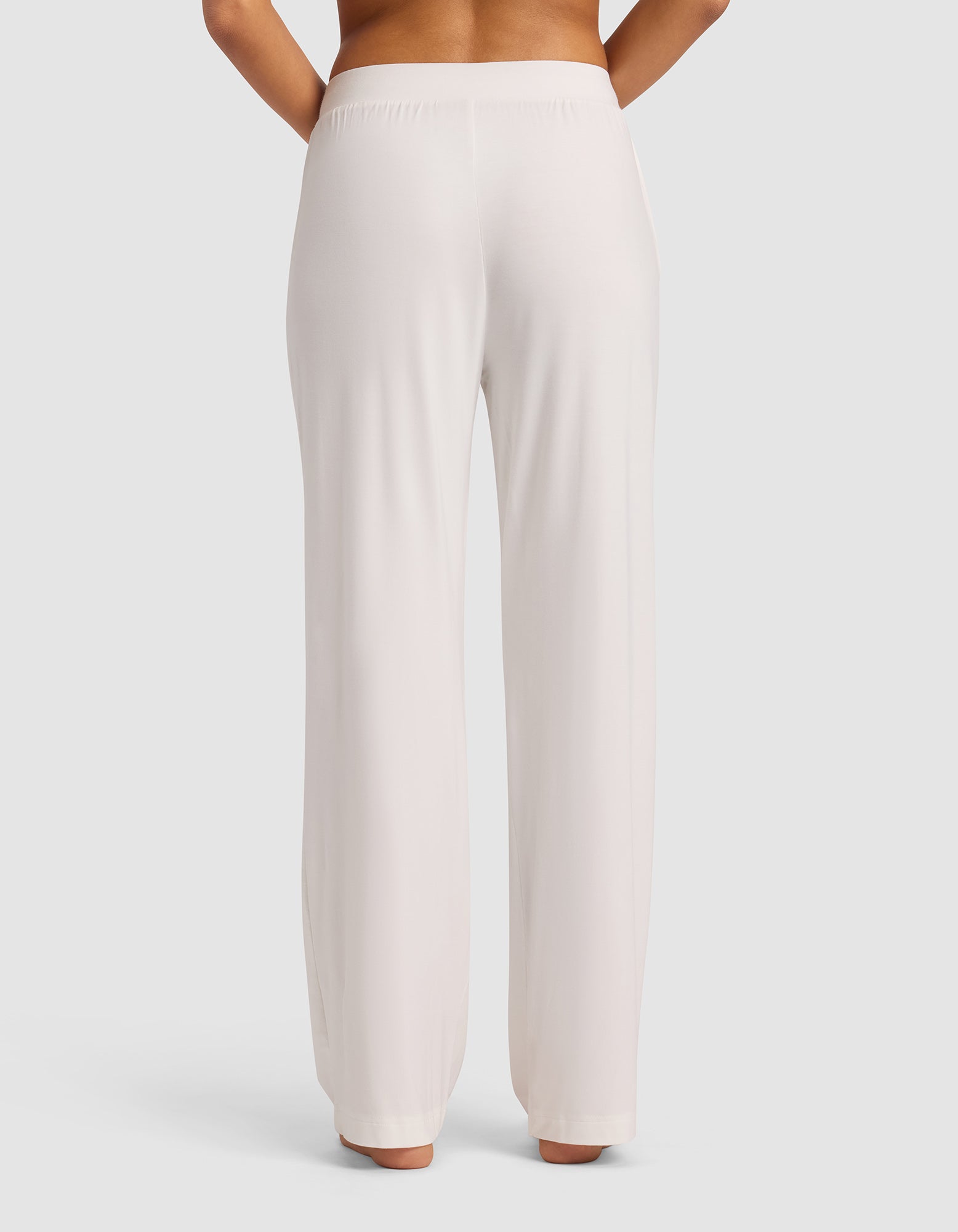 A person stands facing away, wearing Cozy Earth's Women's Bamboo Stretch-Knit Pant in light beige. The high-waisted pants feature a smooth, relaxed fit with no visible pockets or patterns, set against a plain light background.|Color:Ivory
