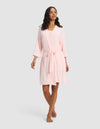 A woman wears the Cozy Earth Women's Bamboo Stretch-Knit Kimono Robe in light pink, knee-length with a belt, standing barefoot against a plain light gray background.|Color:Blossom