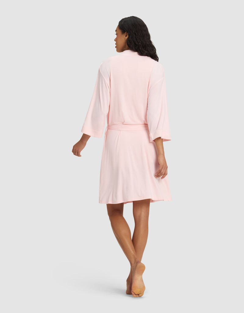 A woman models the Cozy Earth Women's Bamboo Stretch-Knit Kimono Robe in light pink, barefoot on a plain light gray background, shown from the back with her head turned slightly to the side.