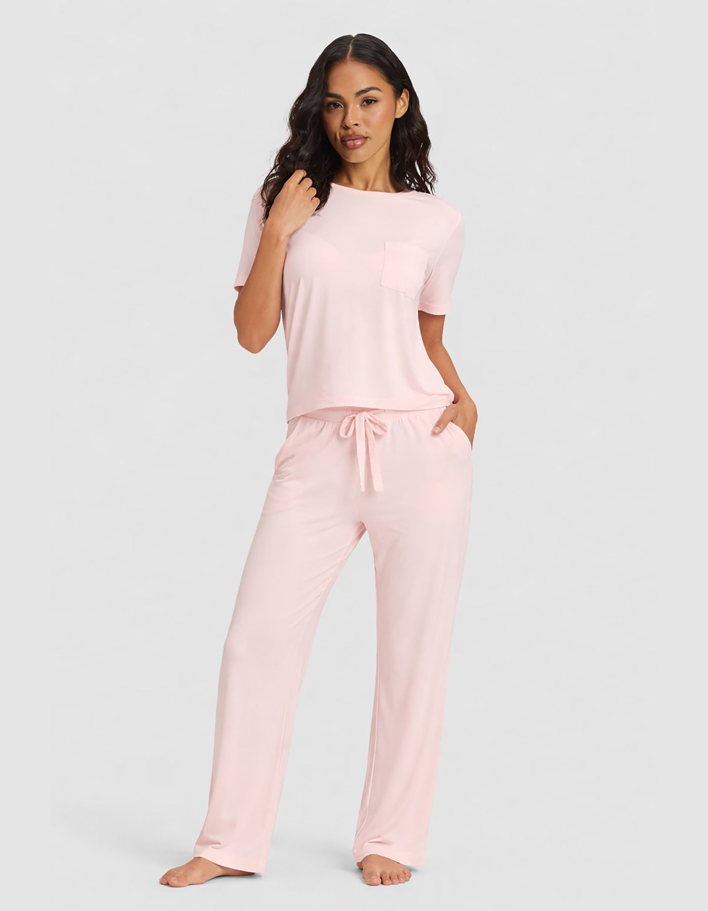 A woman poses barefoot against a plain gray background, wearing Cozy Earth’s Women's Bamboo Stretch-Knit Lounge Tee & Pant Set in light pink with short sleeves, pockets, and drawstring pants.|Color:Blossom