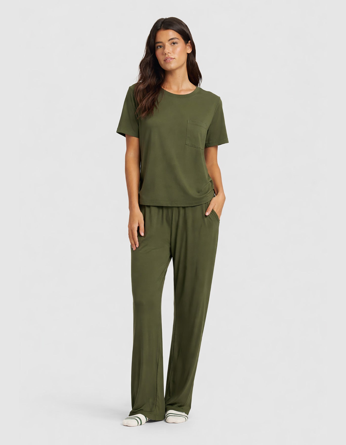 A woman wears the Cozy Earth Women's Bamboo Stretch-Knit Lounge Tee & Pant Set in olive green, featuring a chest pocket and loose pants, accessorized with white sneakers with green stripes.|Color:Olive