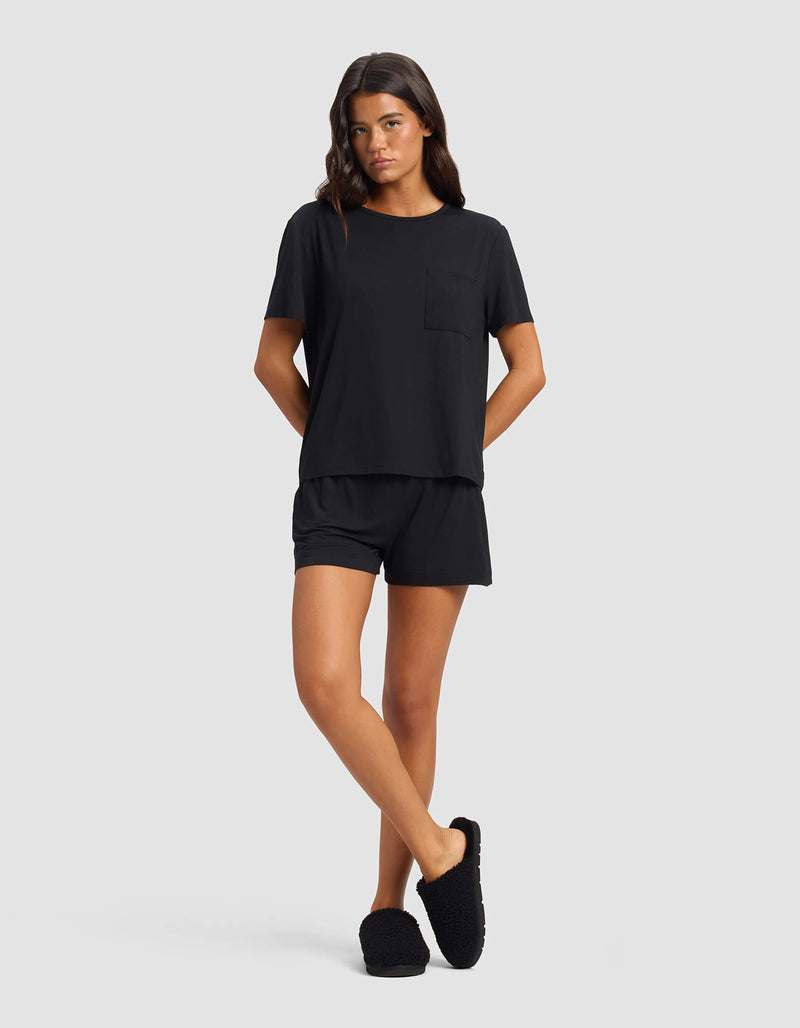 A woman wears the Cozy Earth Women's Bamboo Stretch-Knit Lounge Tee & Short Set in black, featuring a short-sleeve top with chest pocket and matching shorts. She stands against a plain background with hands behind her back and a neutral expression.