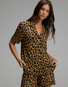 A woman stands against a plain background wearing the Cozy Earth Women’s Bamboo Stretch-Knit Short Sleeve Pajama Top paired with matching shorts, both featuring a bold brown and black leopard print pattern.|Color:Leopard Print in Onyx