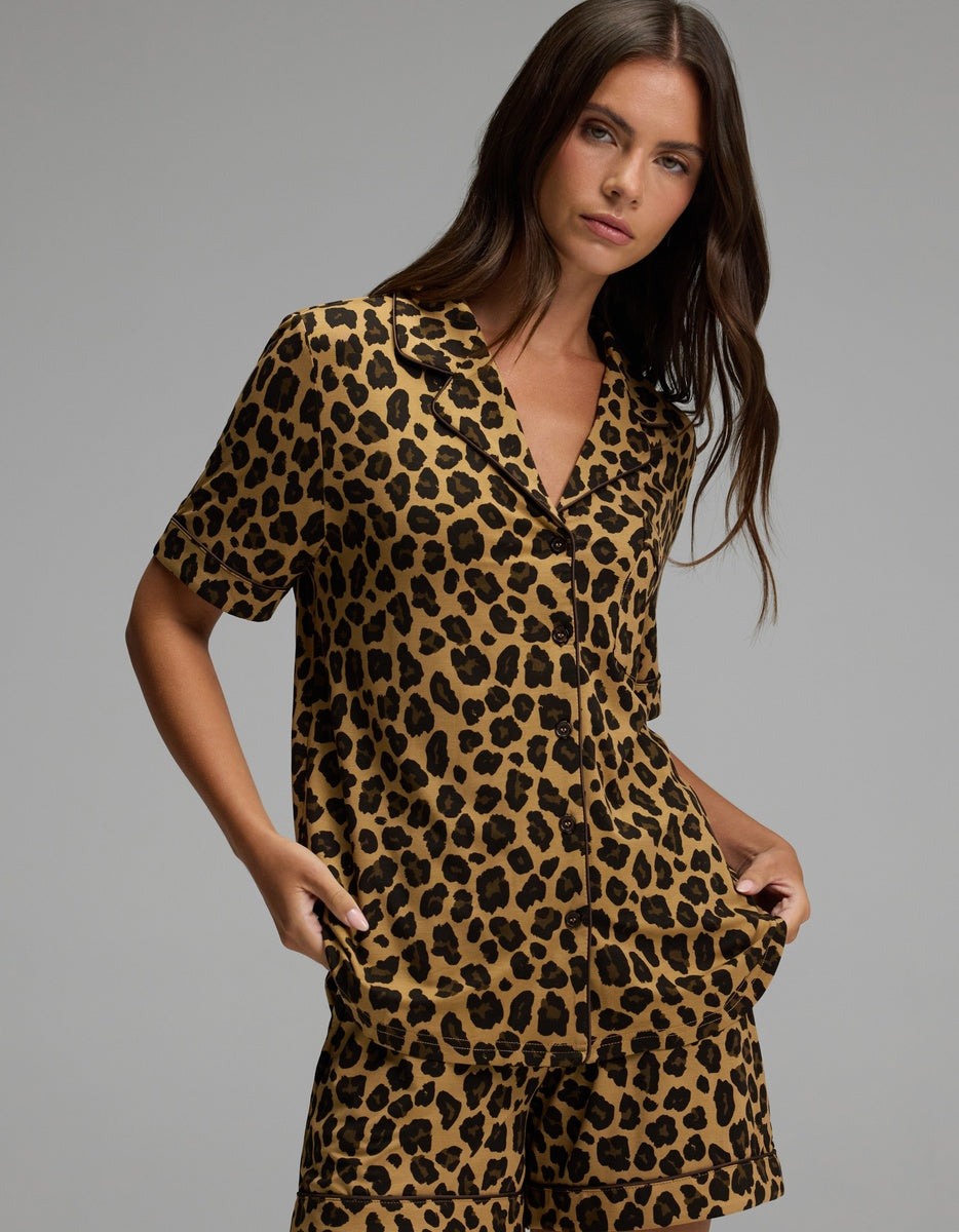 A woman stands against a plain background wearing the Cozy Earth Women’s Bamboo Stretch-Knit Short Sleeve Pajama Top paired with matching shorts, both featuring a bold brown and black leopard print pattern.|Color:Leopard Print in Onyx