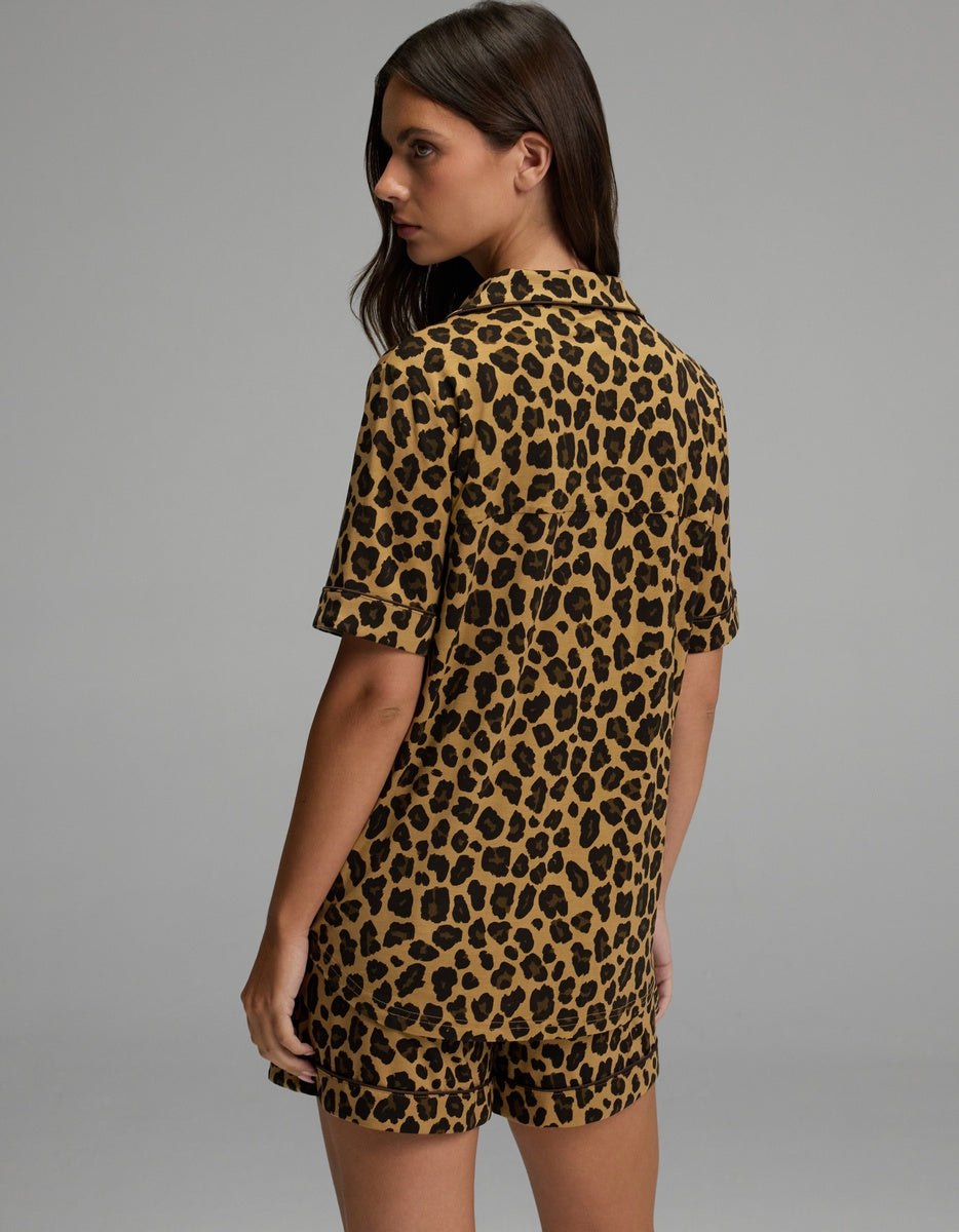 A woman with long brown hair, wearing the Cozy Earth Women’s Bamboo Stretch-Knit Short Sleeve Pajama Top in leopard print and matching shorts, stands turned slightly away from the camera against a plain gray background.