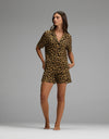 A woman stands barefoot against a plain gray background, wearing the Cozy Earth Women’s Bamboo Stretch-Knit Short Sleeve Pajama Set—featuring shorts, a collared neckline, and a stylish leopard print.|Color:Leopard Print in Onyx