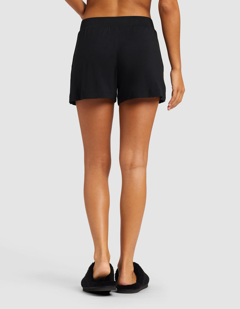A person stands facing away from the camera, wearing Cozy Earth's Women's Bamboo Stretch-Knit Shorts in black. Their arms are relaxed at their sides against a plain light gray background.