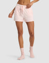 A person models Cozy Earth's Women's Bamboo Stretch-Knit Shorts in light pink, paired with matching socks, standing against a plain light grey background. The image highlights the shorts' fit from lower torso to legs, with one arm bent at the elbow.|Color:Blossom