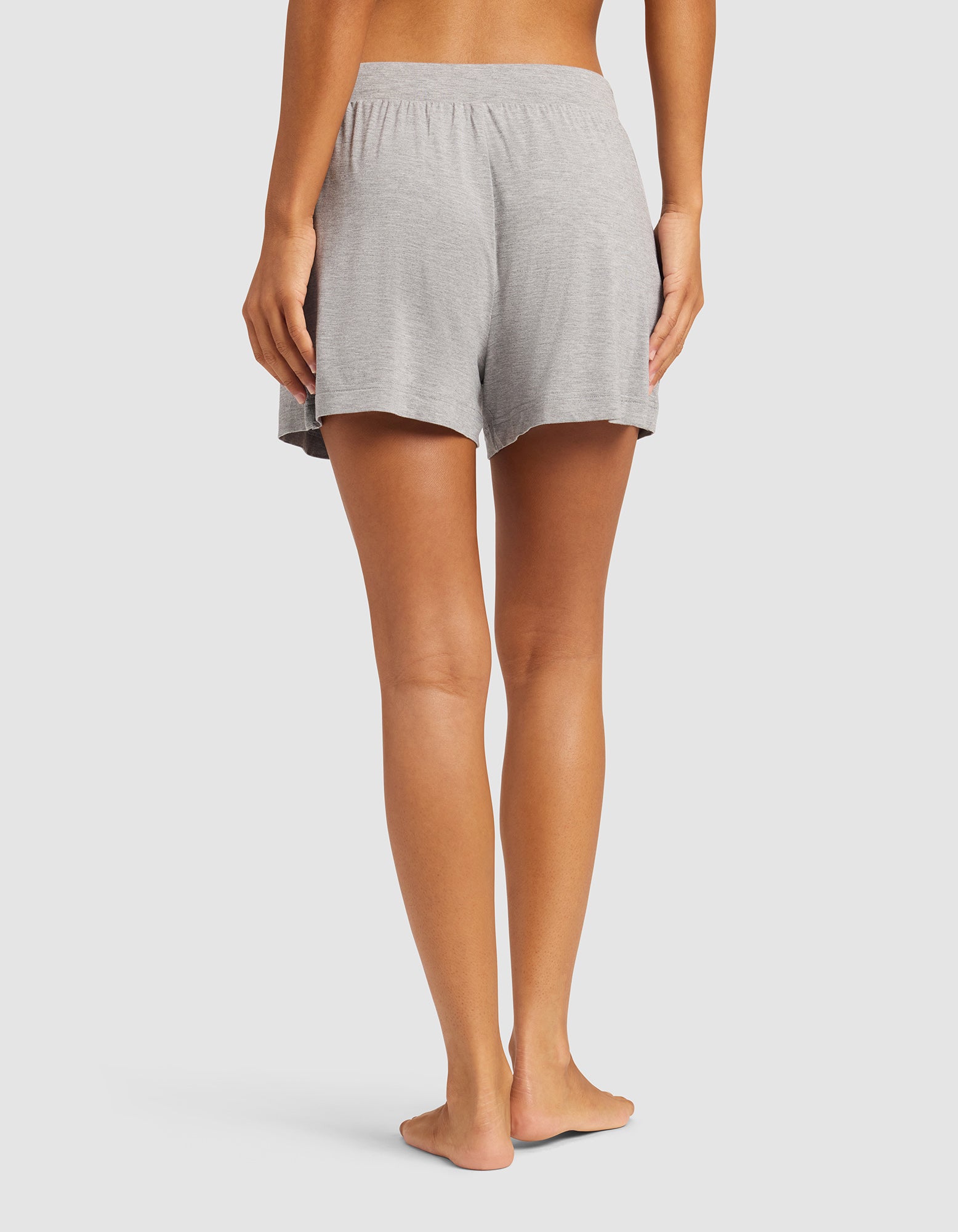 A person stands barefoot on a light background, shown from the waist down and viewed from the back, wearing Cozy Earth Women's Bamboo Stretch-Knit Shorts in light gray.|Color:Heather Grey