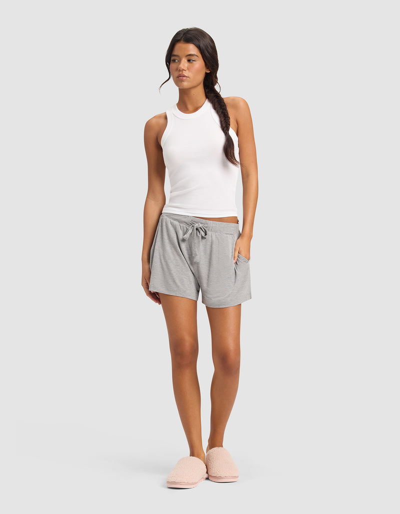 A woman stands against a plain background wearing Cozy Earth's Women's Bamboo Stretch-Knit Shorts in light gray, paired with a white sleeveless tank top and pink fuzzy slippers. Her hair is styled in a loose side braid.