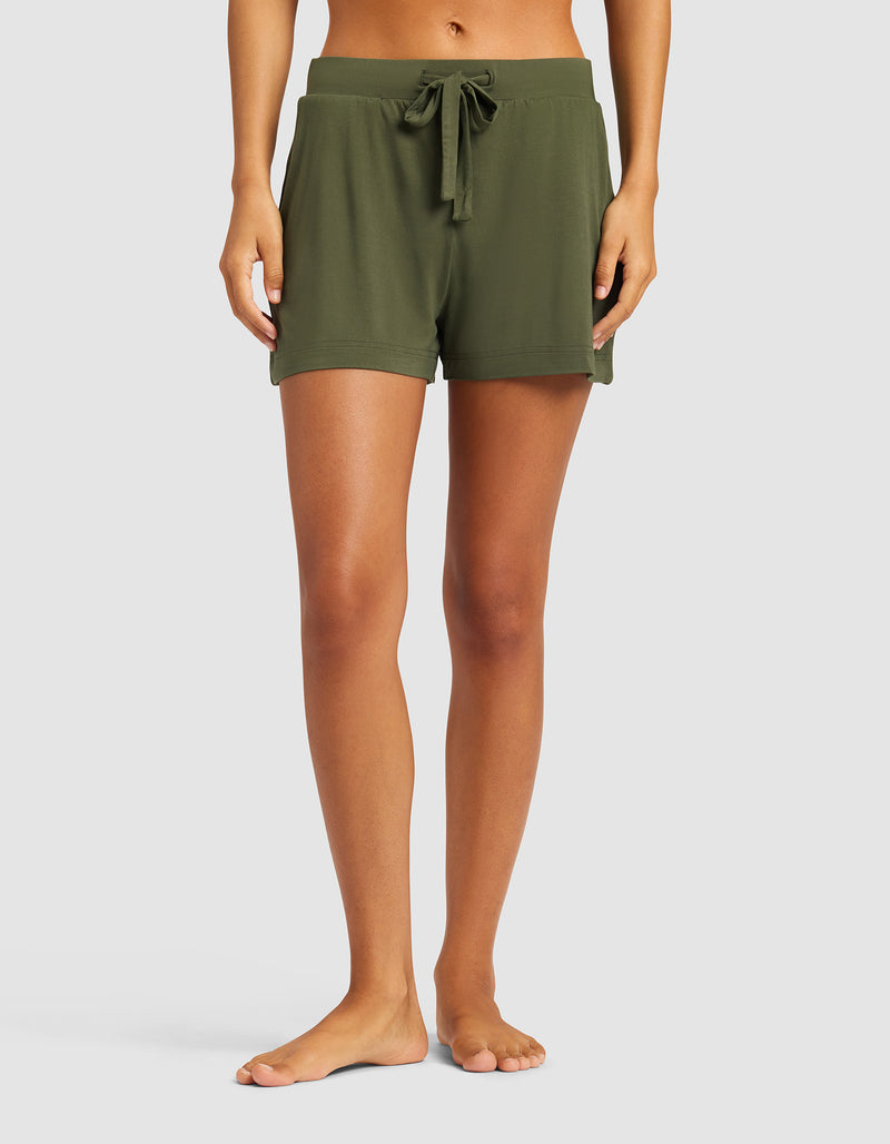 A woman stands barefoot against a plain light background, wearing Cozy Earth's Women's Bamboo Stretch-Knit Shorts in olive green. The shorts feature a drawstring waist, relaxed fit, and side pockets.