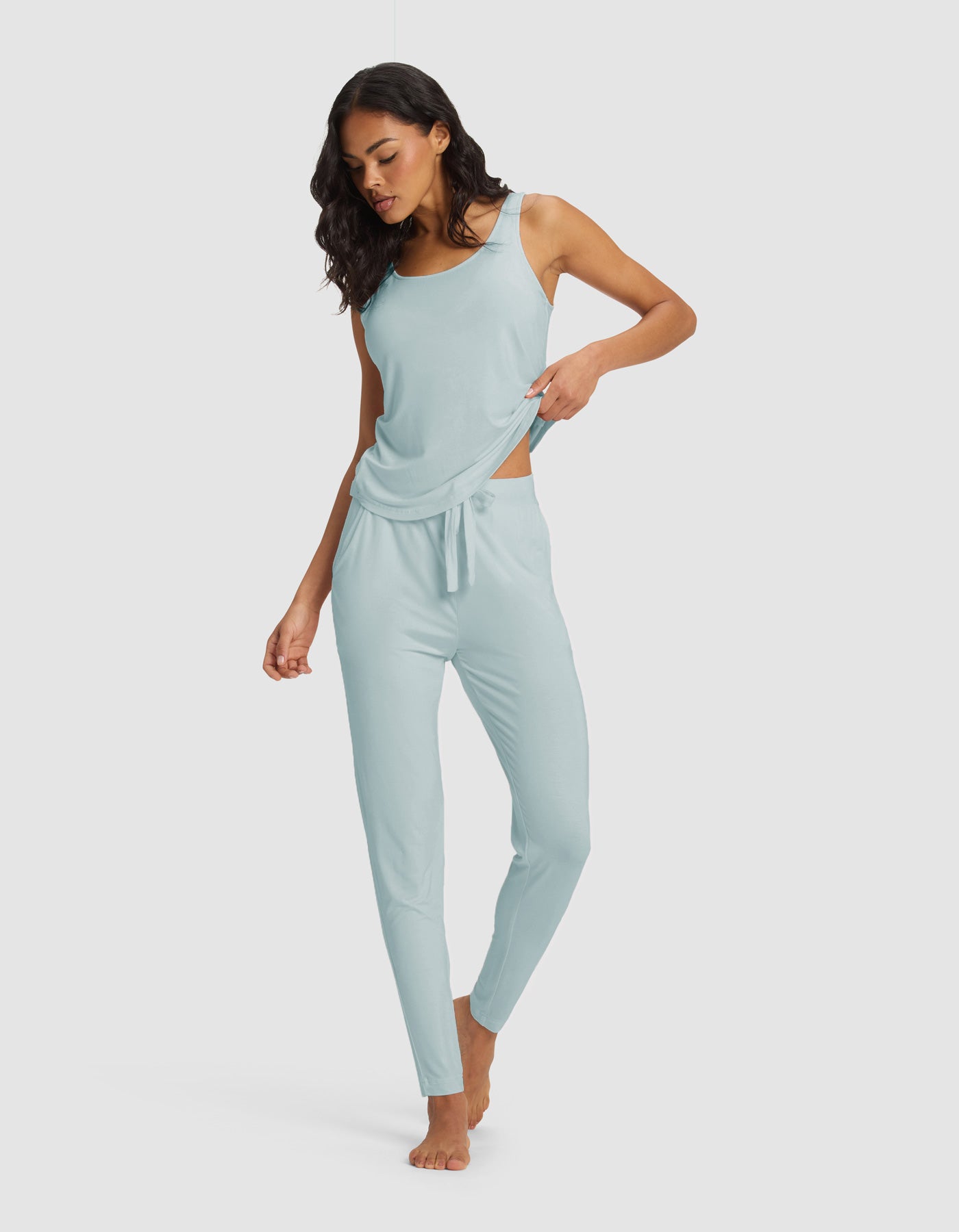 A woman stands barefoot against a plain background, wearing the Cozy Earth Women's Bamboo Stretch-Knit Tank & Tapered Pant Set in light blue, looking down and gently lifting the hem of her top with one hand.|Color:Brook
