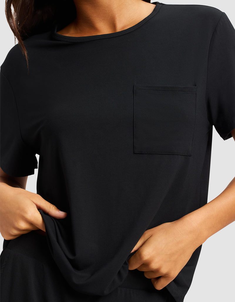 A woman is shown from shoulders to waist wearing the Cozy Earth Women's Bamboo Stretch-Knit Lounge Tee, a plain black short-sleeve shirt with a chest pocket, gently pulling the hem with both hands.