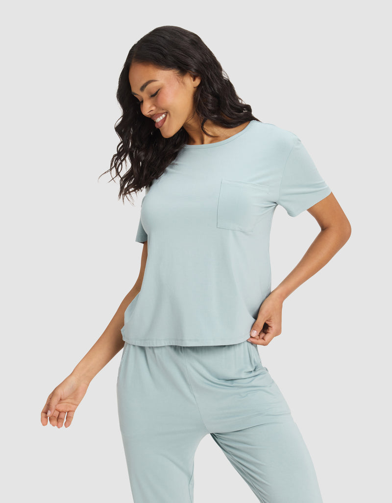 A woman smiles and looks down, wearing the Cozy Earth Women's Bamboo Stretch-Knit Lounge Tee in light blue, featuring short sleeves and a chest pocket, with one hand on her hip against a plain light background.