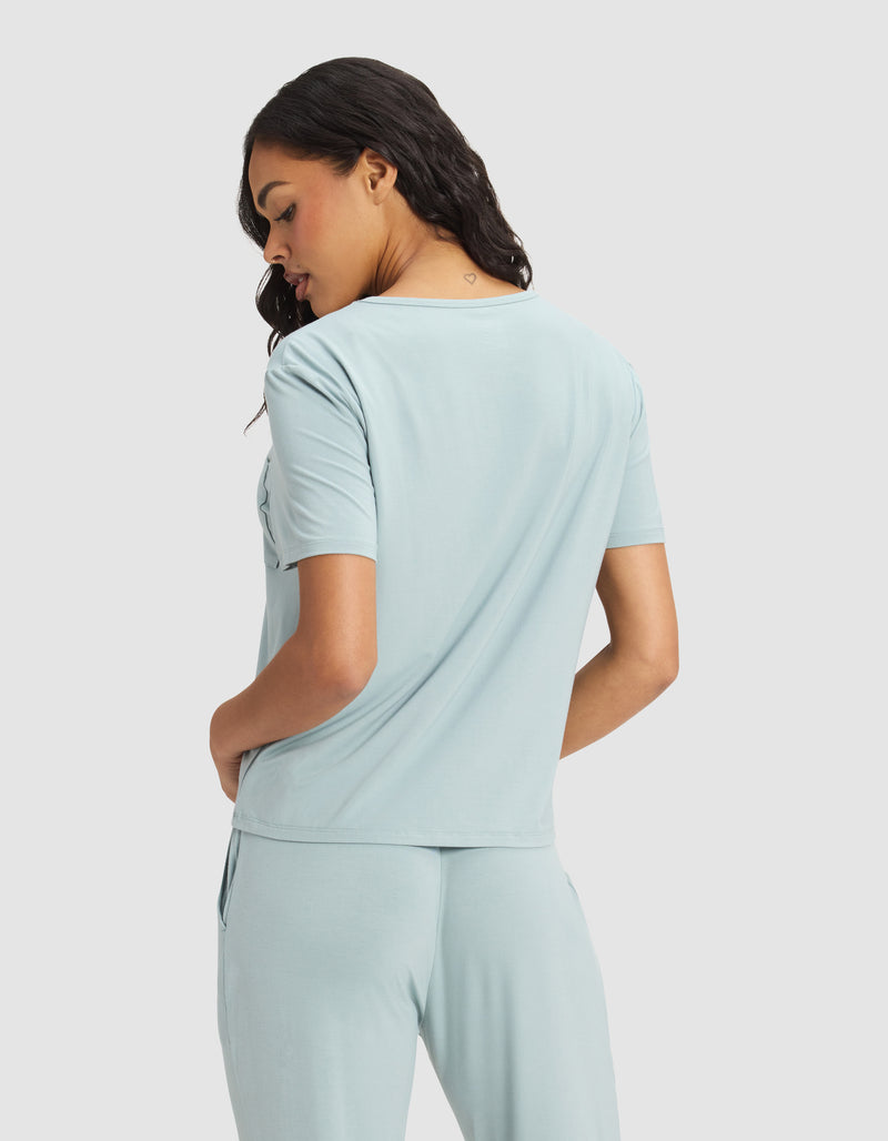 A woman faces away in a Cozy Earth Women's Bamboo Stretch-Knit Lounge Tee, light blue with short sleeves and matching pants. She turns her head slightly, hands in pockets, standing against a plain light background.