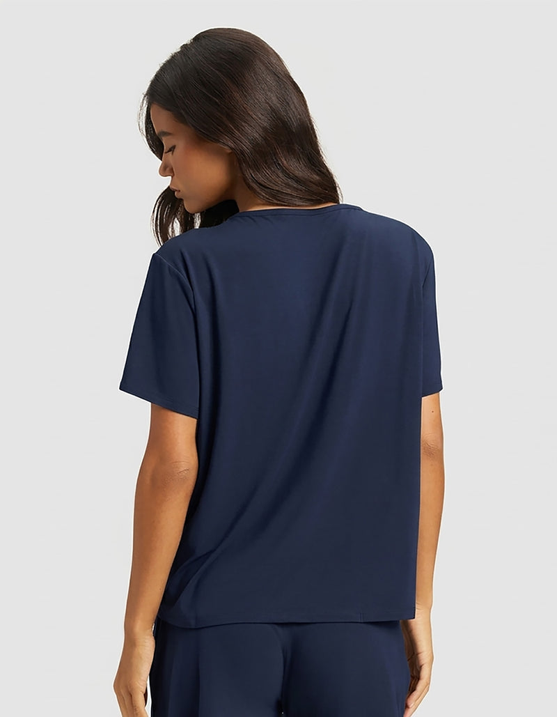 A woman with long dark hair models the Cozy Earth Women's Bamboo Stretch-Knit Lounge Tee & Pant Set in navy, standing with her back to the camera against a plain light gray background.