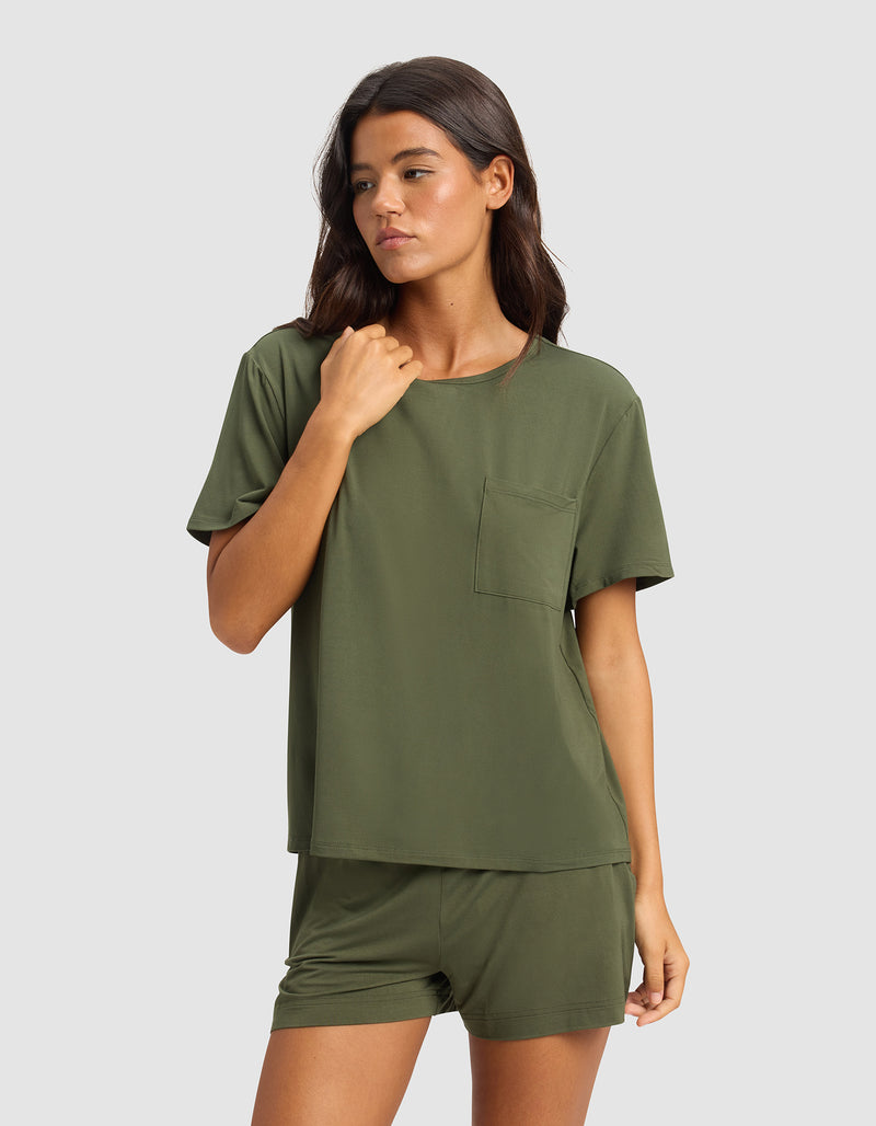 A woman with long brown hair wears the Cozy Earth Women's Bamboo Stretch-Knit Lounge Tee in olive green, paired with matching shorts, posing against a plain light gray background.