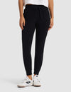A person stands against a light gray background with hands in pockets, wearing Cozy Earth's Women's Brushed Bamboo Jogger Pant in black, paired with a white shirt and white sneakers.|Color:Black