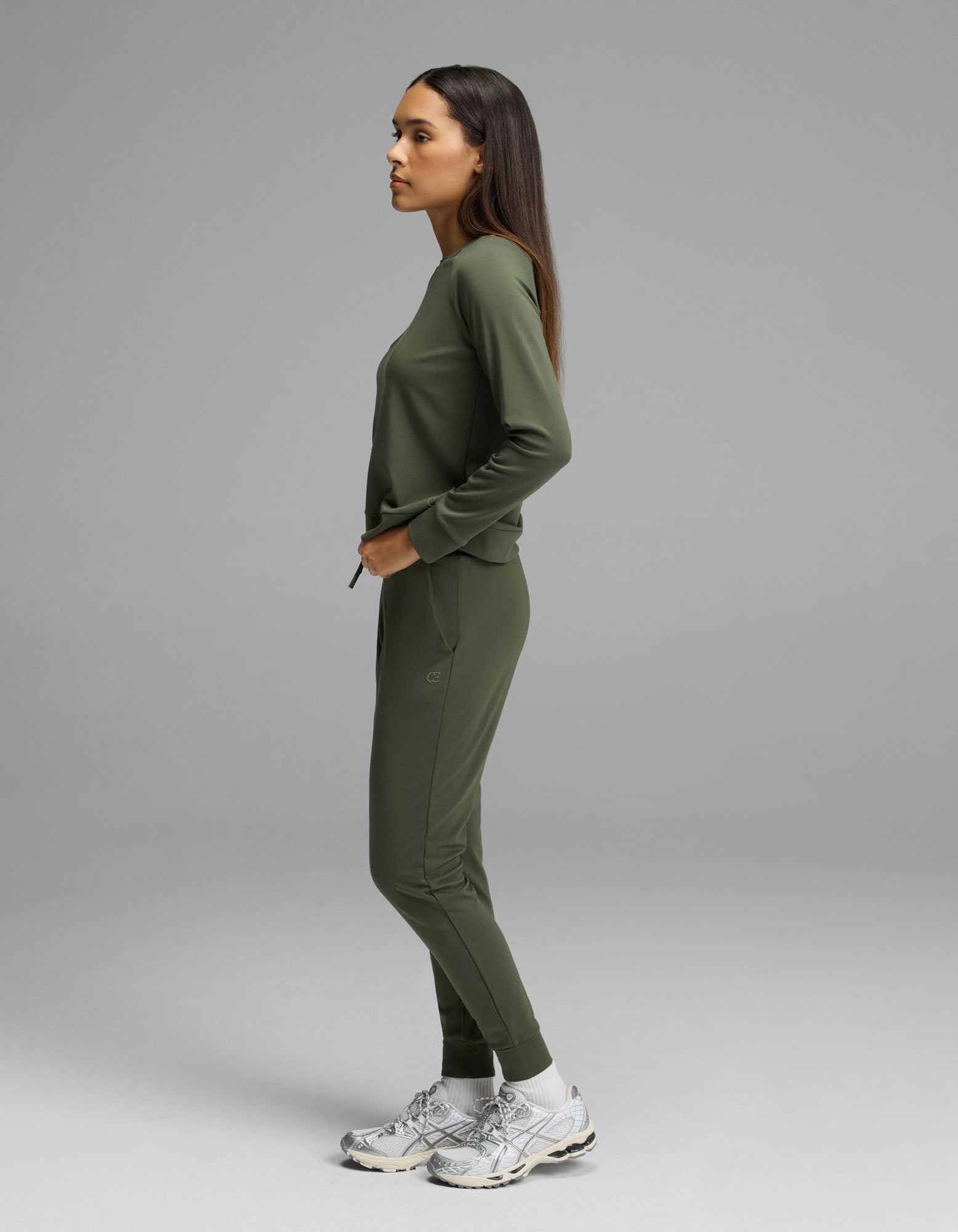 A person stands in profile against a plain background, wearing the Women’s Brushed Bamboo Jogger Set by Cozy Earth with white sneakers.|Color:Olive