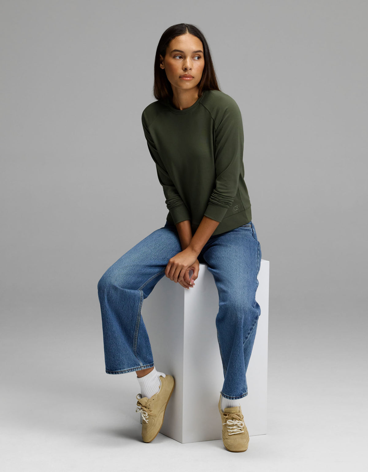 A woman wears the Cozy Earth Women’s Brushed Bamboo Pullover Crew in green and blue jeans as she sits on a white block against a plain gray background, looking slightly to her left. She pairs her outfit with beige sneakers and white socks.|Color:Olive