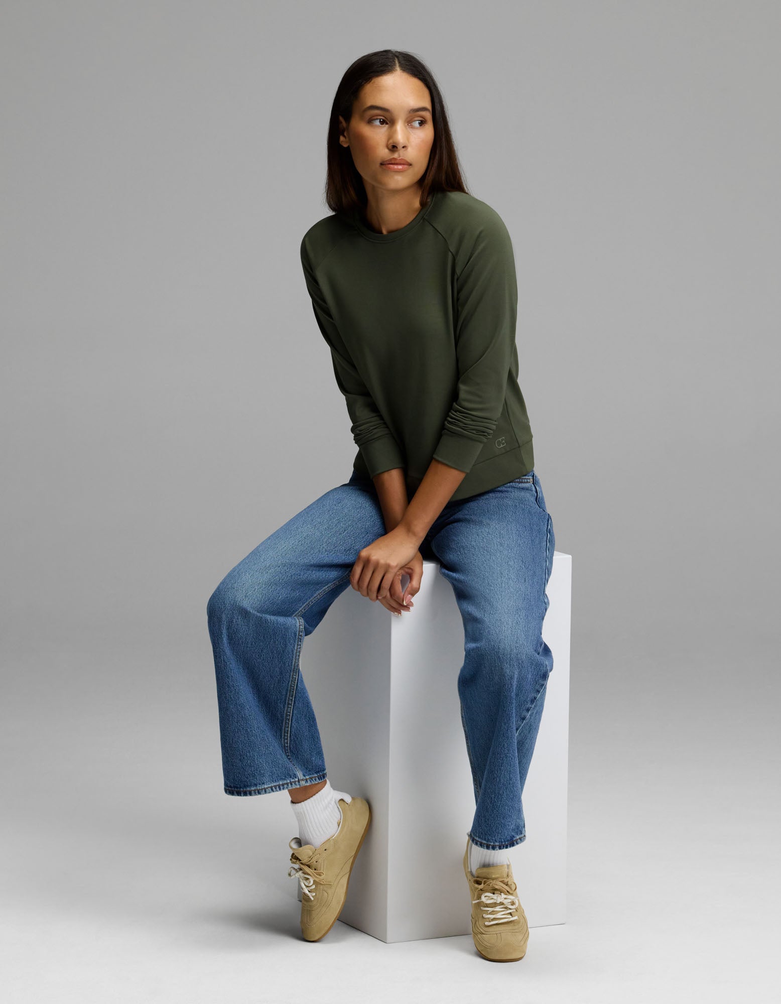 A woman wears the Cozy Earth Women’s Brushed Bamboo Pullover Crew in green and blue jeans as she sits on a white block against a plain gray background, looking slightly to her left. She pairs her outfit with beige sneakers and white socks.