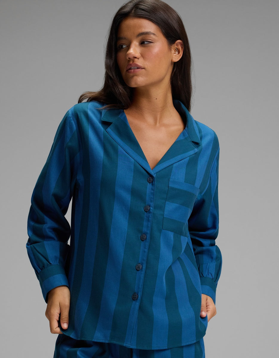 A woman wearing the Cozy Earth Women’s Belmont Long Sleeve Pajama Top in a teal and blue striped pattern stands against a plain gray background, looking to her left with one hand in her pocket.