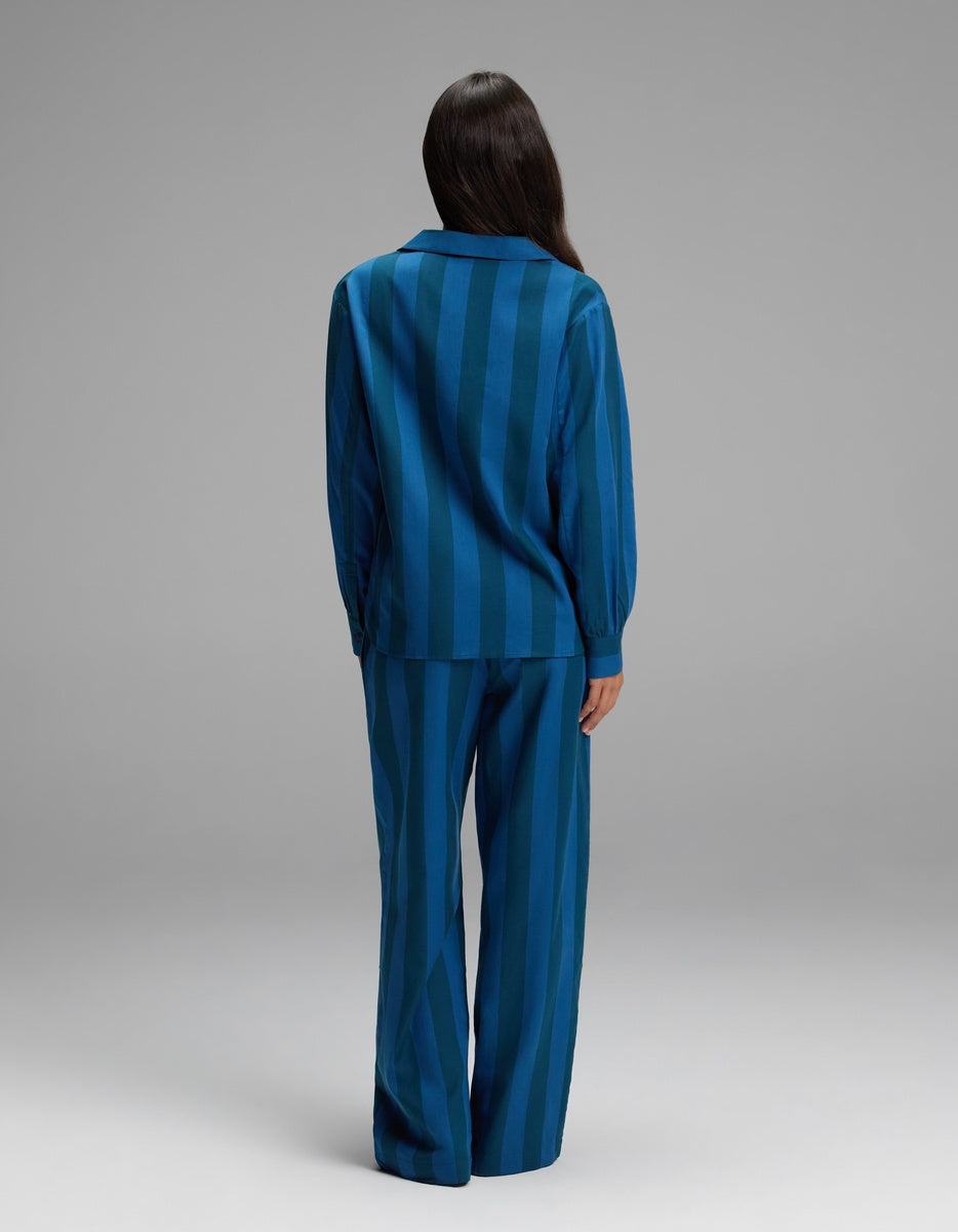 A person stands facing away, wearing the Cozy Earth Women’s Belmont Long Sleeve Pajama Set in a relaxed blue striped design with long sleeves and wide-leg pants. The background is plain gray.