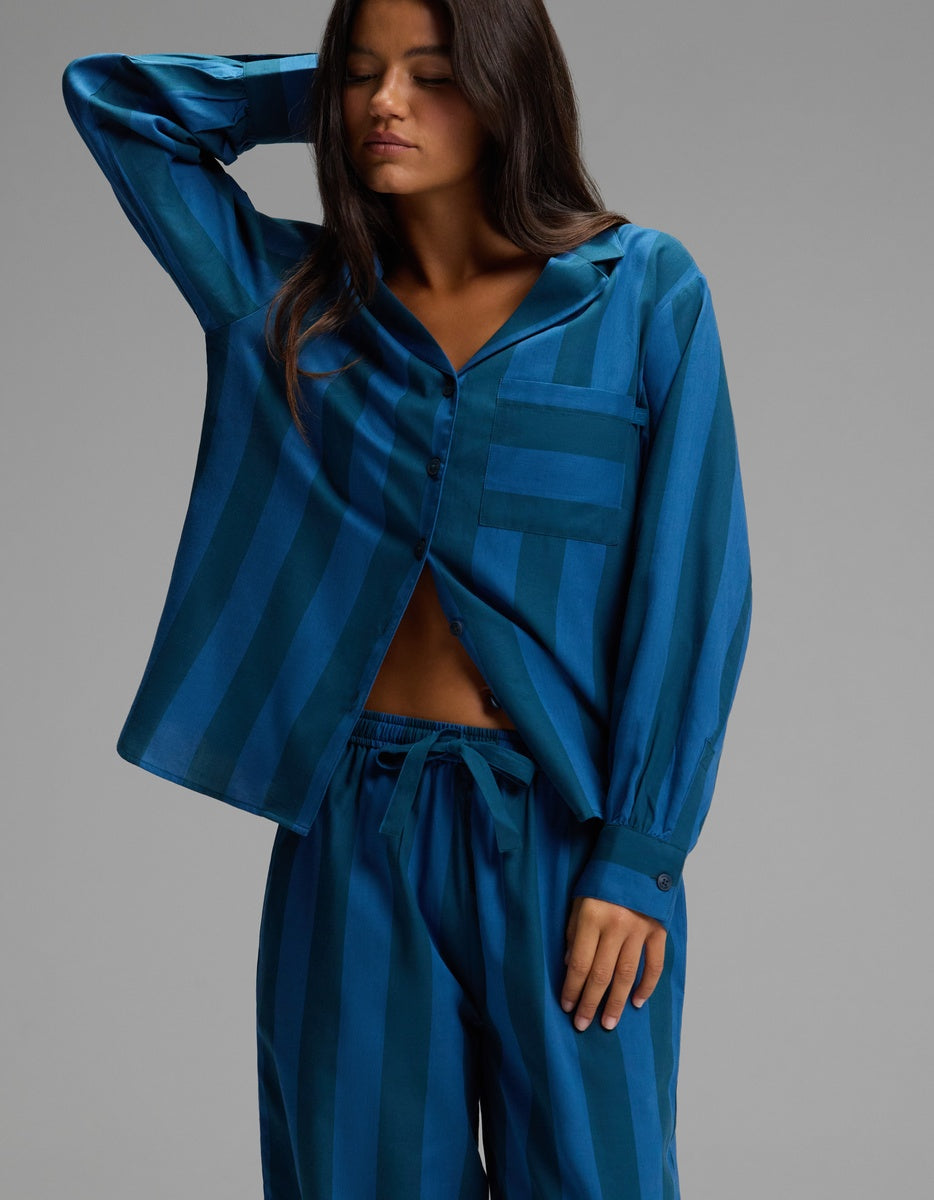 A woman wearing the Cozy Earth Women’s Belmont Long Sleeve Pajama Set in a blue striped print stands with one hand behind her head, looking downward against a plain gray background.