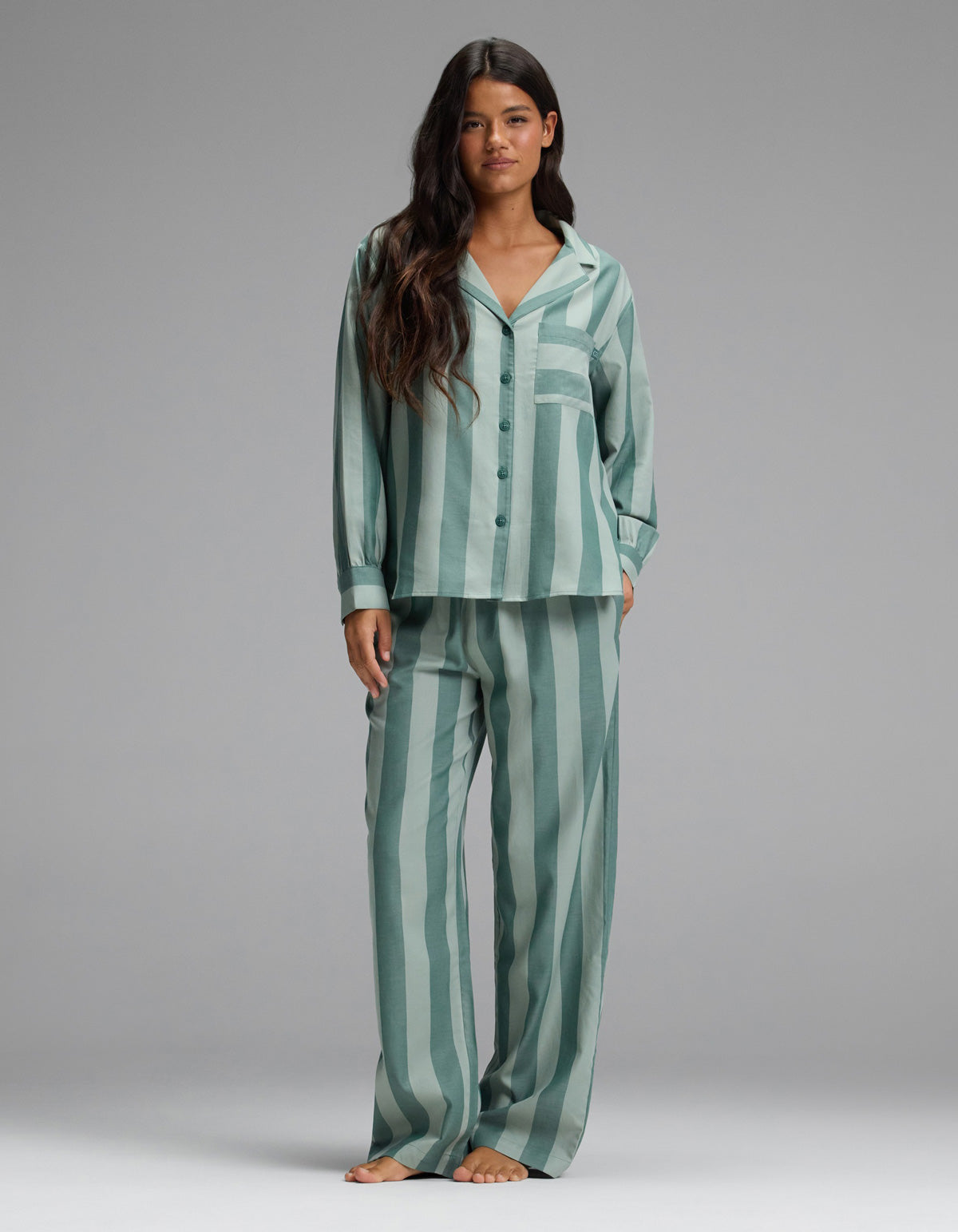 A woman stands barefoot against a plain gray background, wearing the Cozy Earth Women’s Belmont Long Sleeve Pajama Set in Rosemary Stripe—featuring loose-fitting, vertically striped green and light green button-up top with matching pants.