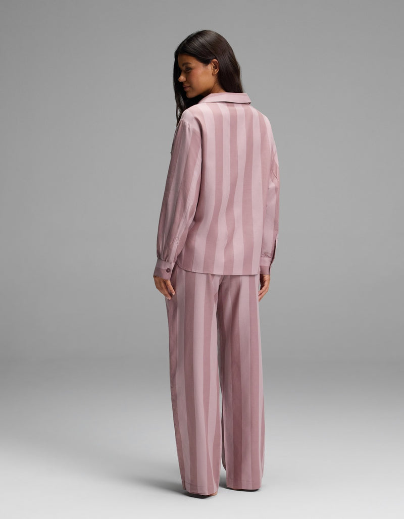 A woman stands with her back to the camera, wearing the Cozy Earth Women’s Belmont Long Sleeve Pajama Set—a loose-fitting, pink striped shirt with long sleeves and matching wide-leg pants—set against a plain gray background.