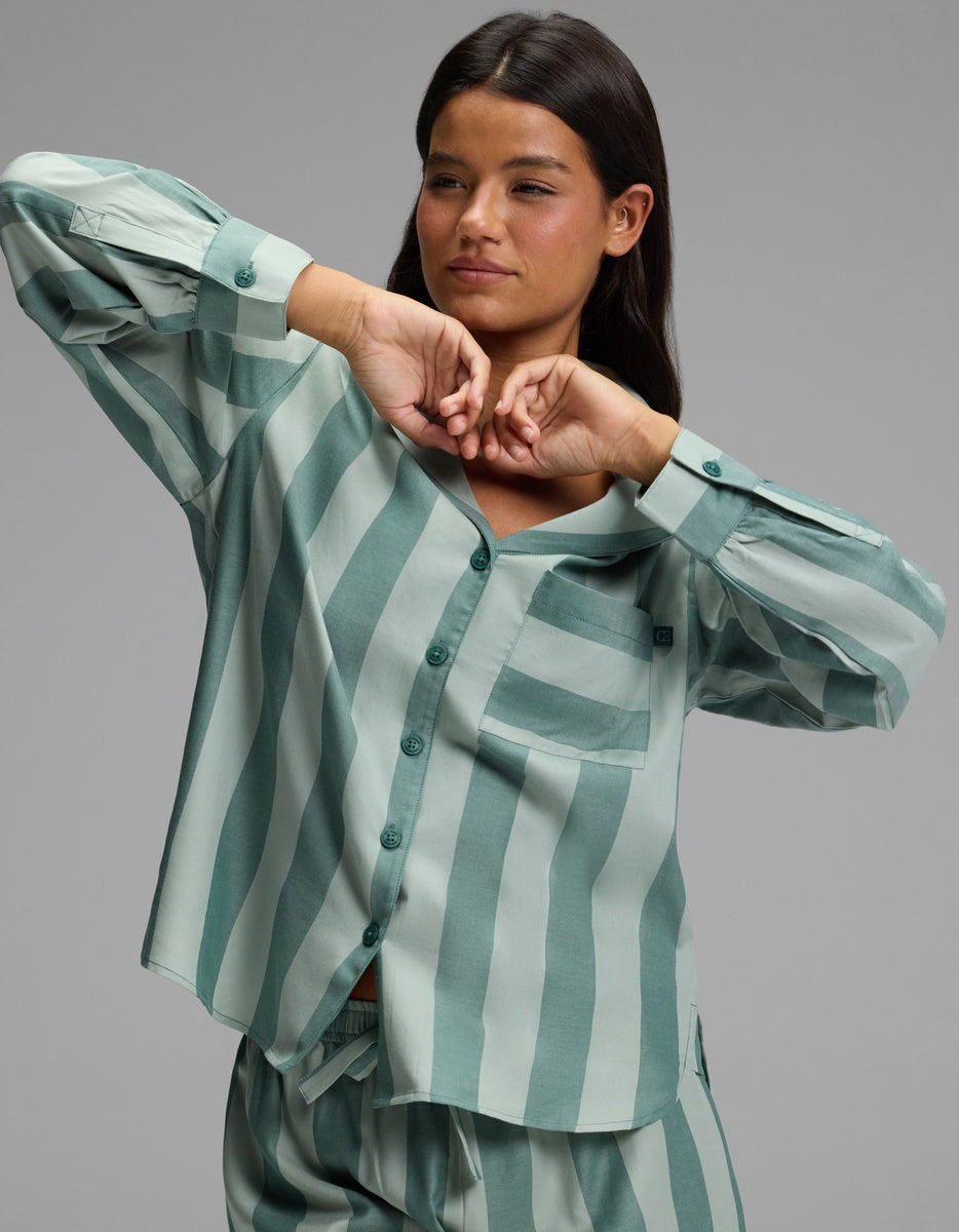 A woman wearing the Cozy Earth Women’s Belmont Long Sleeve Pajama Top in a loose, green and light green vertically striped pattern, along with matching pants, poses with her hands raised near her face against a plain gray background.