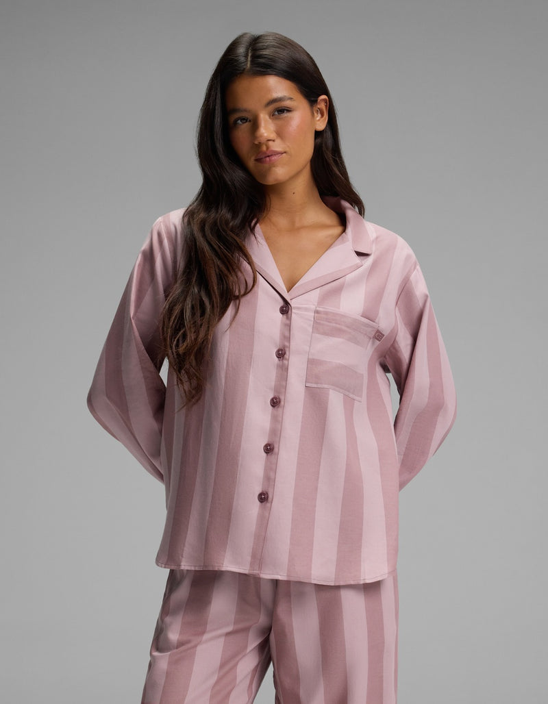 A woman with long dark hair wears the Cozy Earth Women’s Belmont Long Sleeve Pajama Top—a matching pink and mauve striped button-up pajama top with a chest pocket—while standing against a plain gray background.