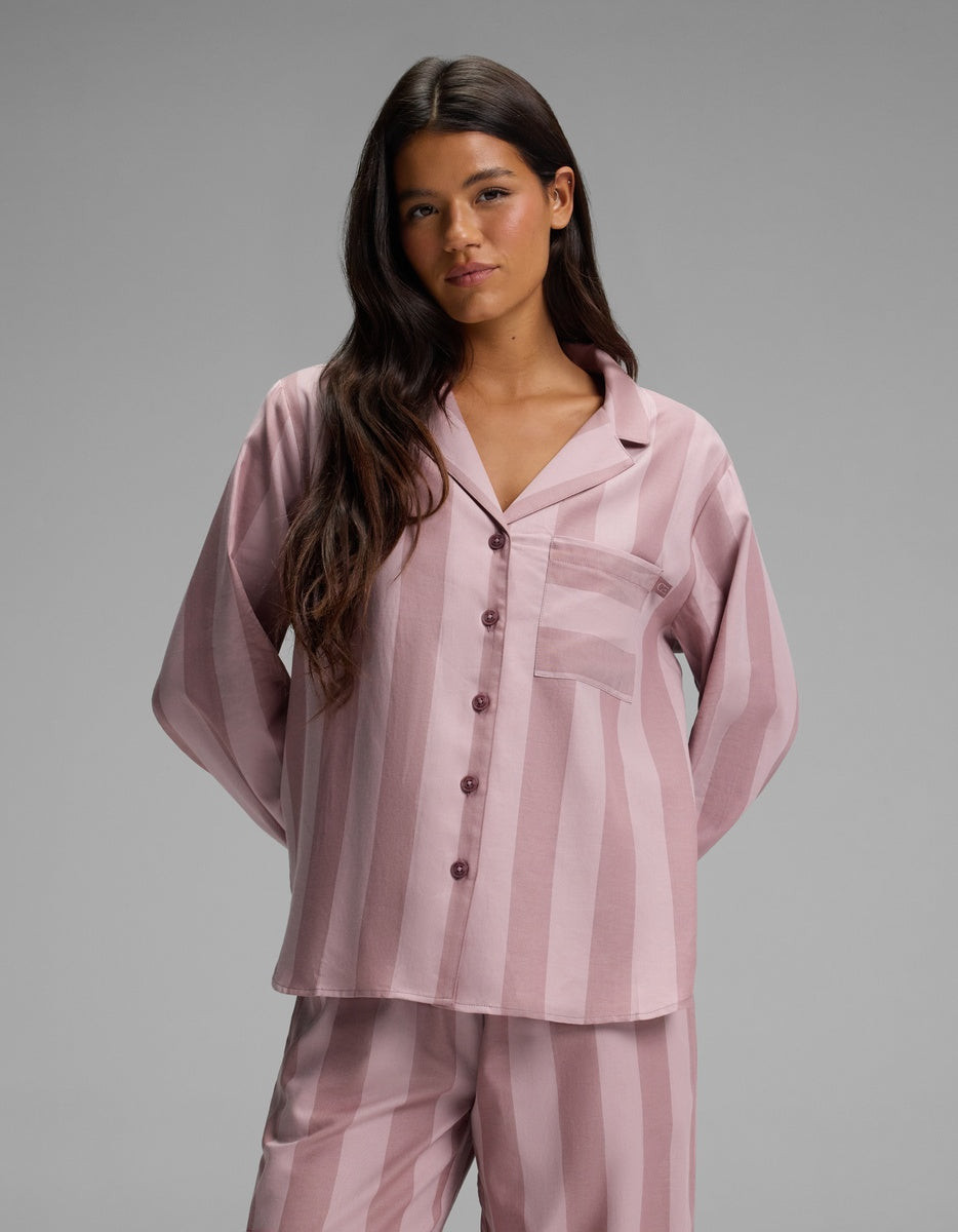 A woman with long dark hair wears the Cozy Earth Women’s Belmont Long Sleeve Pajama Top—a matching pink and mauve striped button-up pajama top with a chest pocket—while standing against a plain gray background.|Color:Viola Stripe